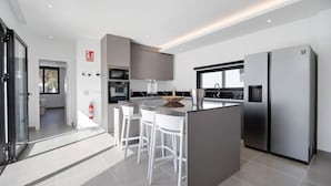 Une cuisine moderne dotée d’un îlot central, de tabourets de bar blancs, d’appareils électroménagers en acier inoxydable et offrant une vue sur l’extérieur.