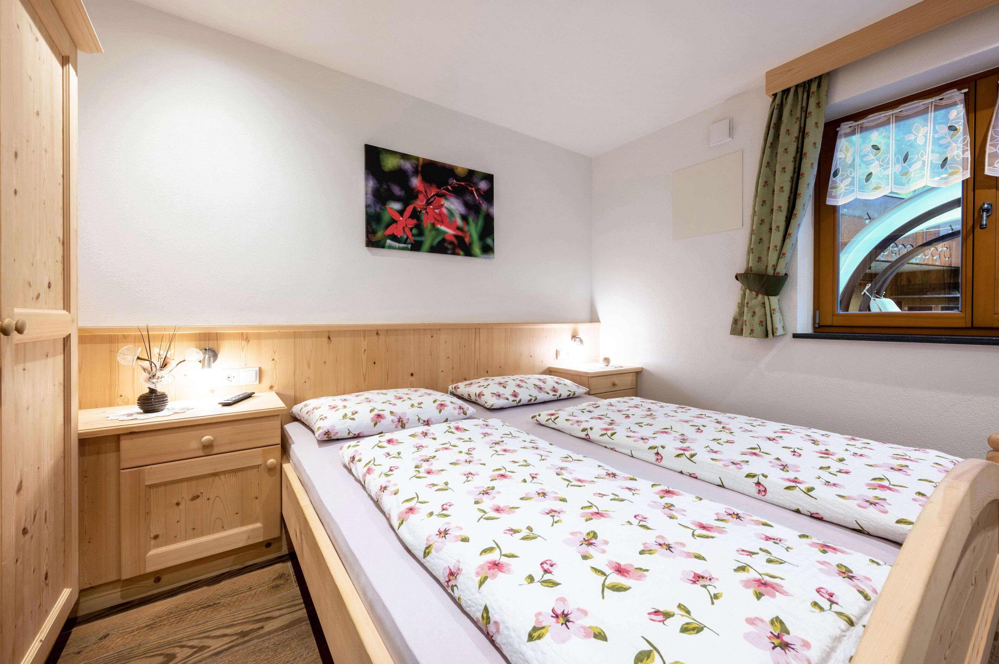 Una habitación con cabecera de madera, cama con ropa de cama floral, mesita de noche con lámpara y ventana con cortinas.