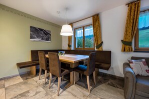 Un'area pranzo con tavolo in legno, sedie imbottite, una panca, un lampadario e un quadro appeso al muro. Una finestra con tende completa l'ambiente.