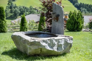 Una fuente de piedra con un caño, rodeada de césped y árboles, con una casa y una cerca al fondo.