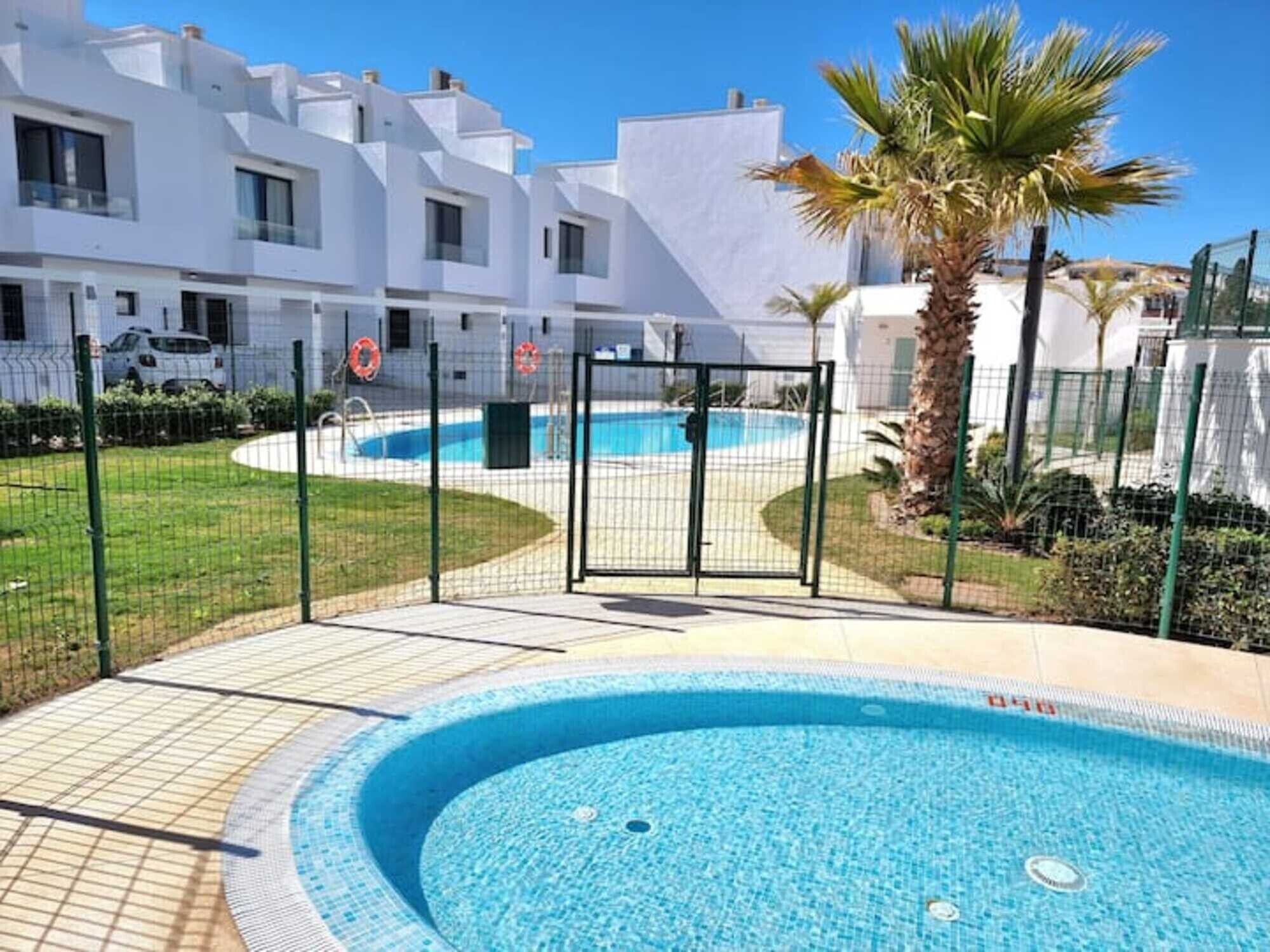 Una piscina rodeada por una cerca, con una palmera y un edificio blanco al fondo.
