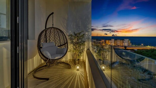 Un balcón con una silla colgante, plantas en macetas y vista al mar y edificios al atardecer.
