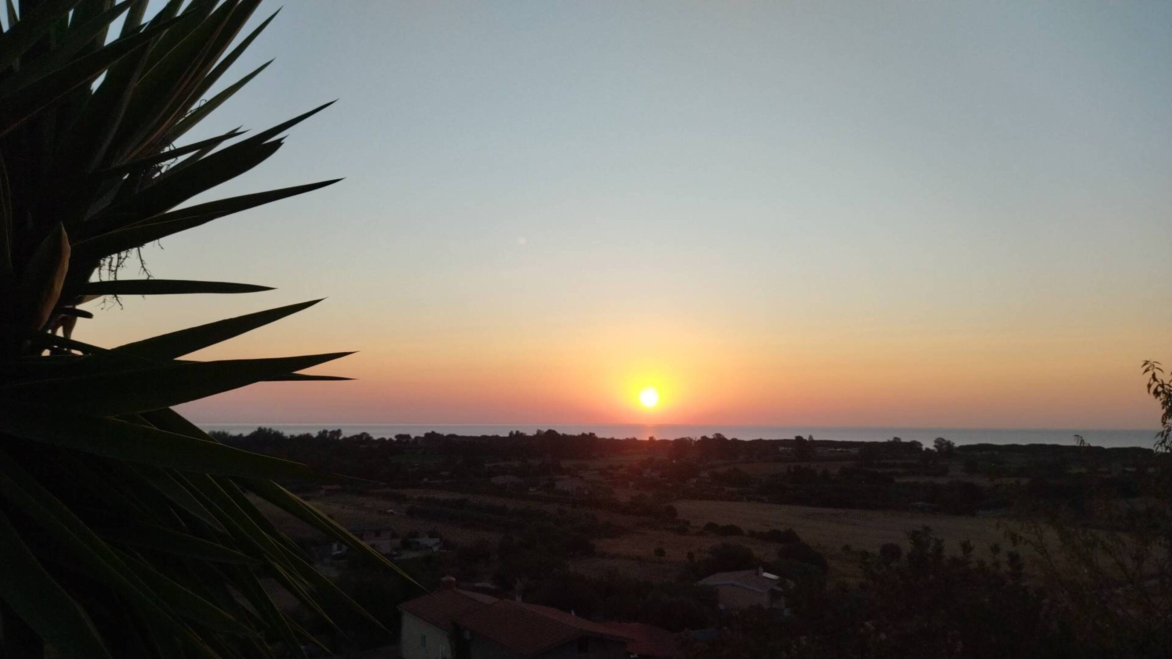 Auringonlasku maaseutumaisemassa, jossa näkyy taloja ja kasvillisuutta.