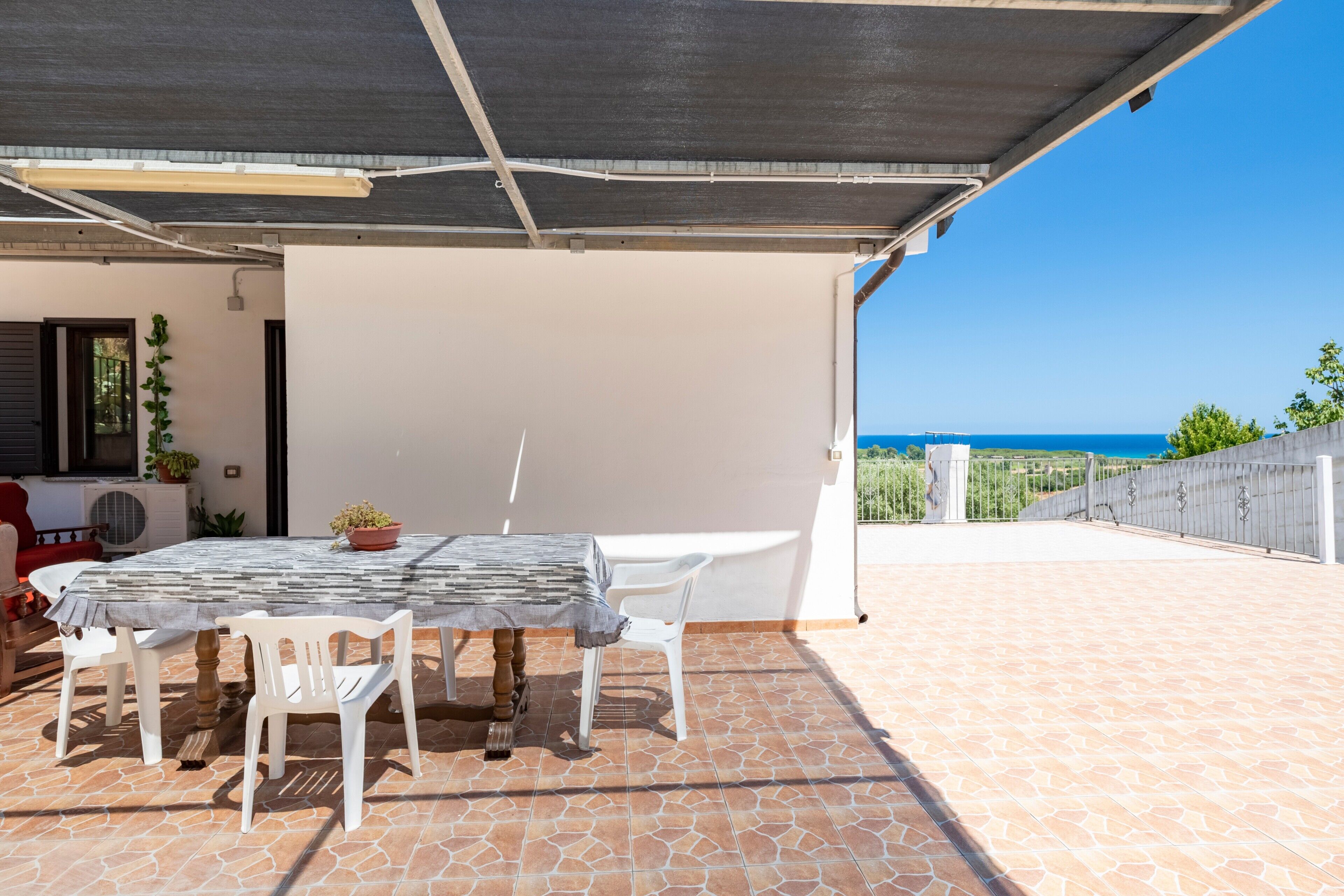 A covered patio with a table and chairs, a potted plant, and a view of the sea.