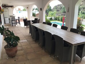 Een terras buiten met een lange tafel en stoelen, een potplant en uitzicht op een zwembad.