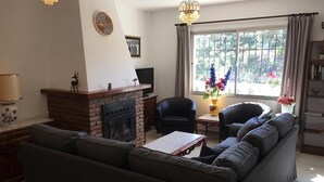 Un salon comprenant un canapé, une cheminée, deux fauteuils, une table avec un livre et une fenêtre avec des rideaux.
