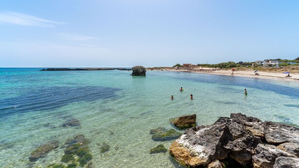 Escena costera de playa con agua cristalina, rocas y edificios a lo lejos.