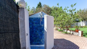 Un pequeño ducha al aire libre, con paredes de azulejos azules, exterior blanco y un sendero de ladrillo que conduce a ella.