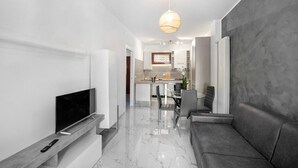 Un salón moderno con un televisor de pantalla plana, un sofá, un comedor con mesa y sillas, y una cocina al fondo.