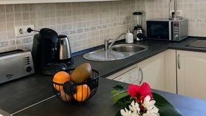 Une cuisine avec des meubles blancs, un grille-pain, une machine à café, un four à micro-ondes, un évier et un panier de fruits contenant des oranges et une fleur.