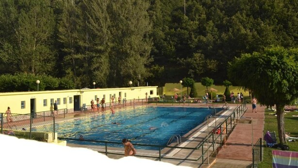 A swimming pool with people swimming and walking around. There is a building with a yellow exterior and a tree-lined hill in the background.