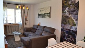A living room with a brown sectional sofa, a glass coffee table, a painting of a waterfall, and a striped tablecloth on a dining table.