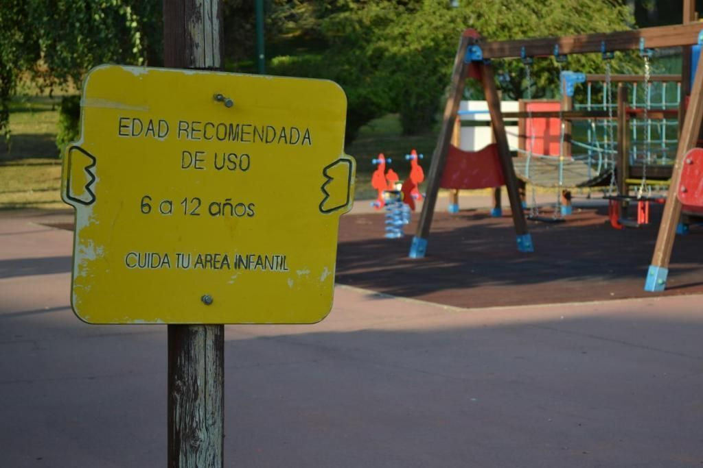 A yellow sign with text in Spanish, placed outdoors with playground equipment in the background.