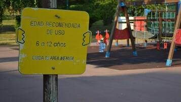 A yellow sign with text in Spanish, placed outdoors with playground equipment in the background.