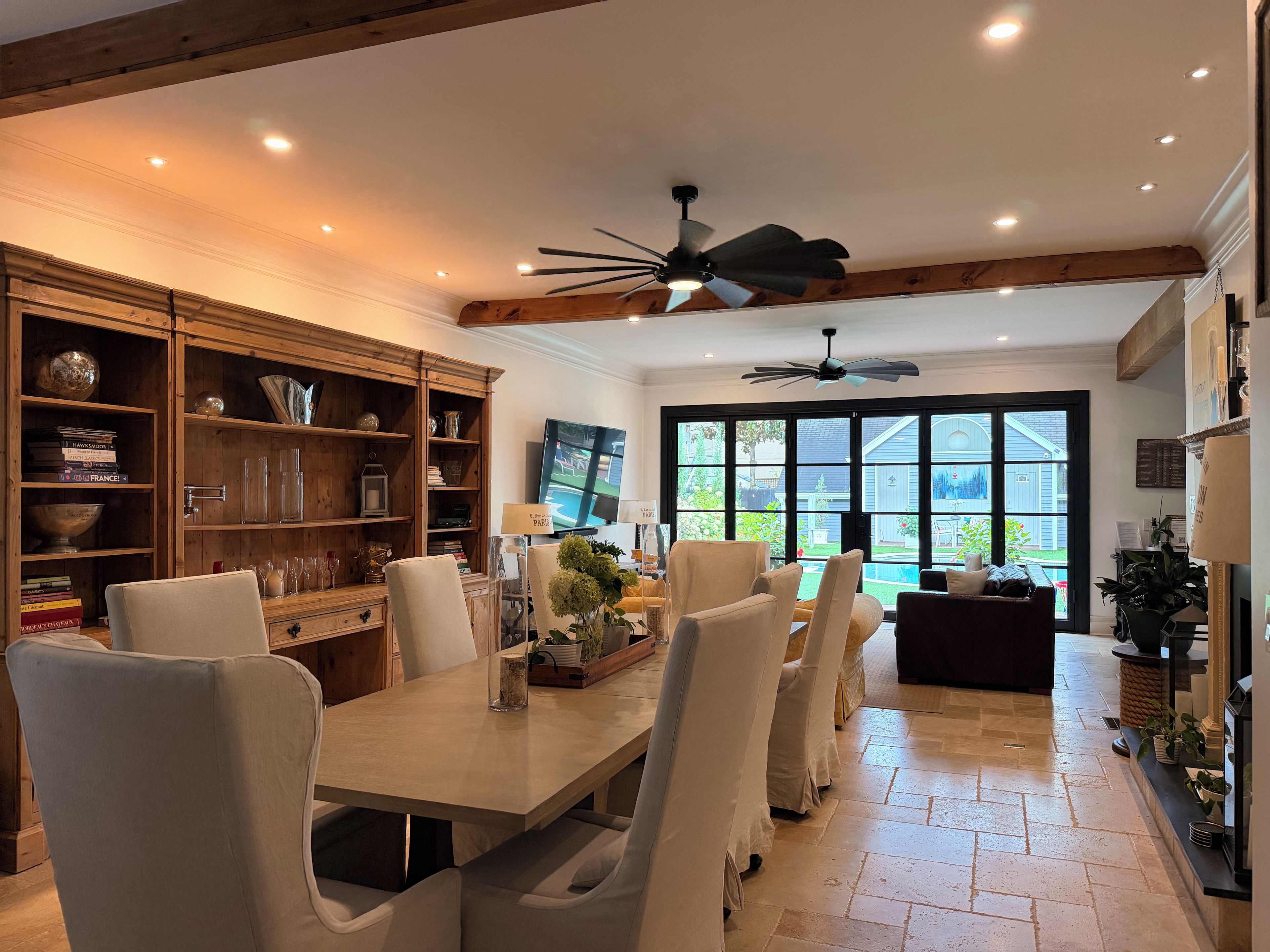 A dining area with a large table, chairs, a ceiling fan, and a view of the outdoors through a large window.