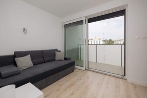 Un salon moderne avec un canapé gris, une petite table blanche et une porte-fenêtre coulissante donnant sur un balcon.