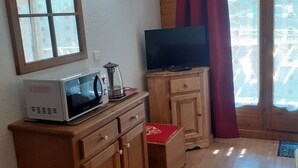 Un espace de vie compact comprenant un four à micro-ondes, un téléviseur posé sur un meuble en bois et une fenêtre agrémentée de rideaux à motifs.