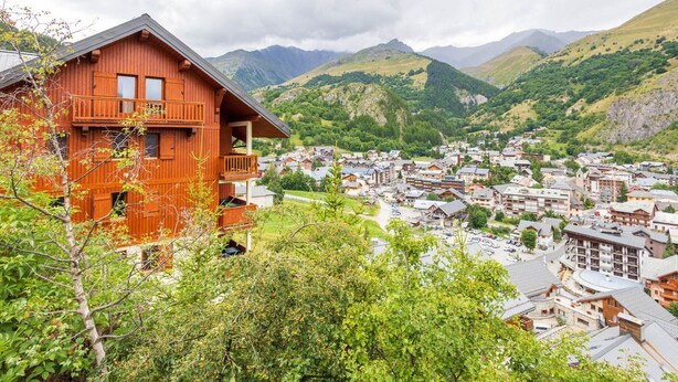 Un chalet en bois doté d’un balcon, situé dans un cadre pittoresque, avec un village de montagne et une végétation luxuriante en arrière-plan.