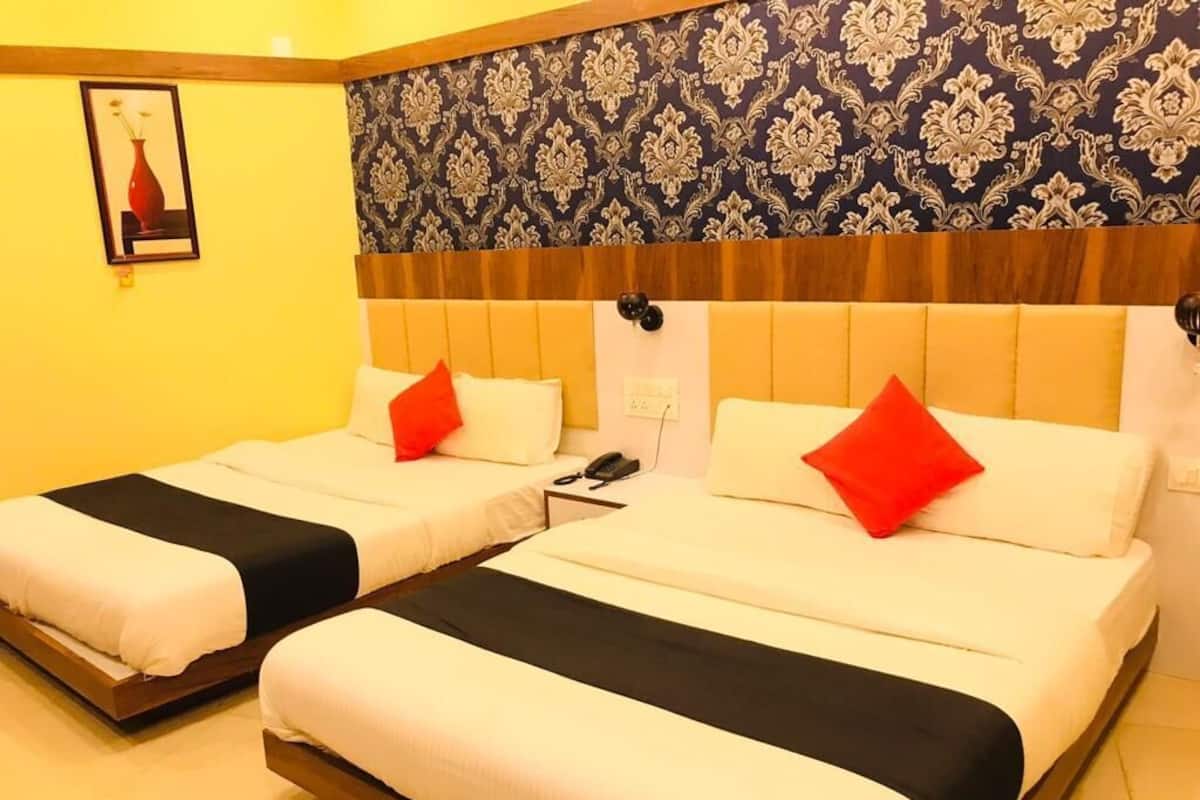 Quarto de hotel com duas camas, cada uma com roupa de cama branca e almofadas vermelhas. O quarto tem papel de parede estampado e cabeceira de madeira.