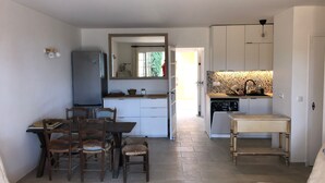 Cocina compacta con refrigerador, estufa y lavaplatos. Cuenta con una zona de comedor con mesa y sillas, y un pequeño rincón de descanso con un banco.