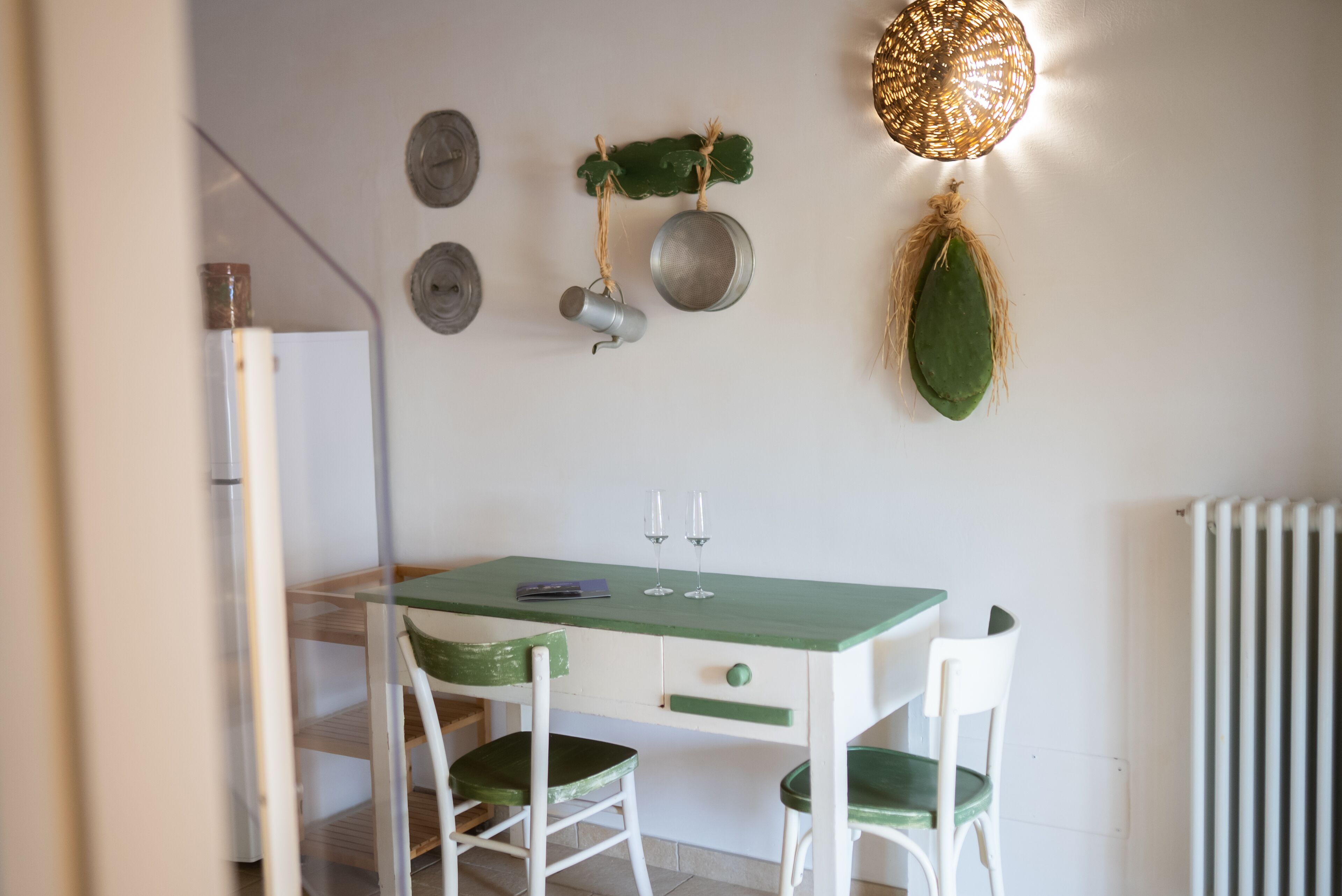A dining area with a green table and white chairs, a hanging light fixture, and wall decorations including a leaf and metal pots.