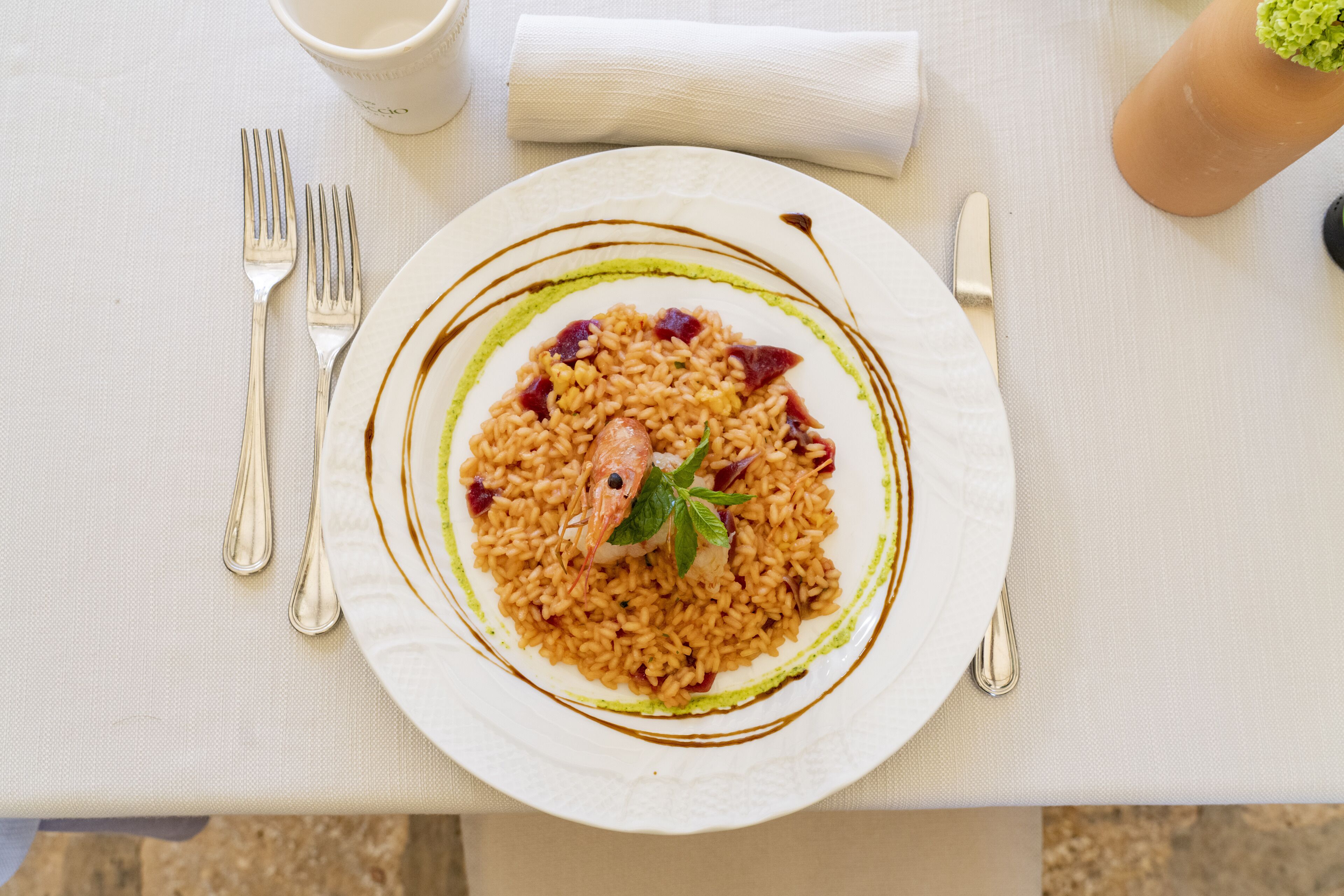 Un plato de risotto con una guarnición de camarón, acompañado de un tenedor y un cuchillo sobre un mantel blanco.
