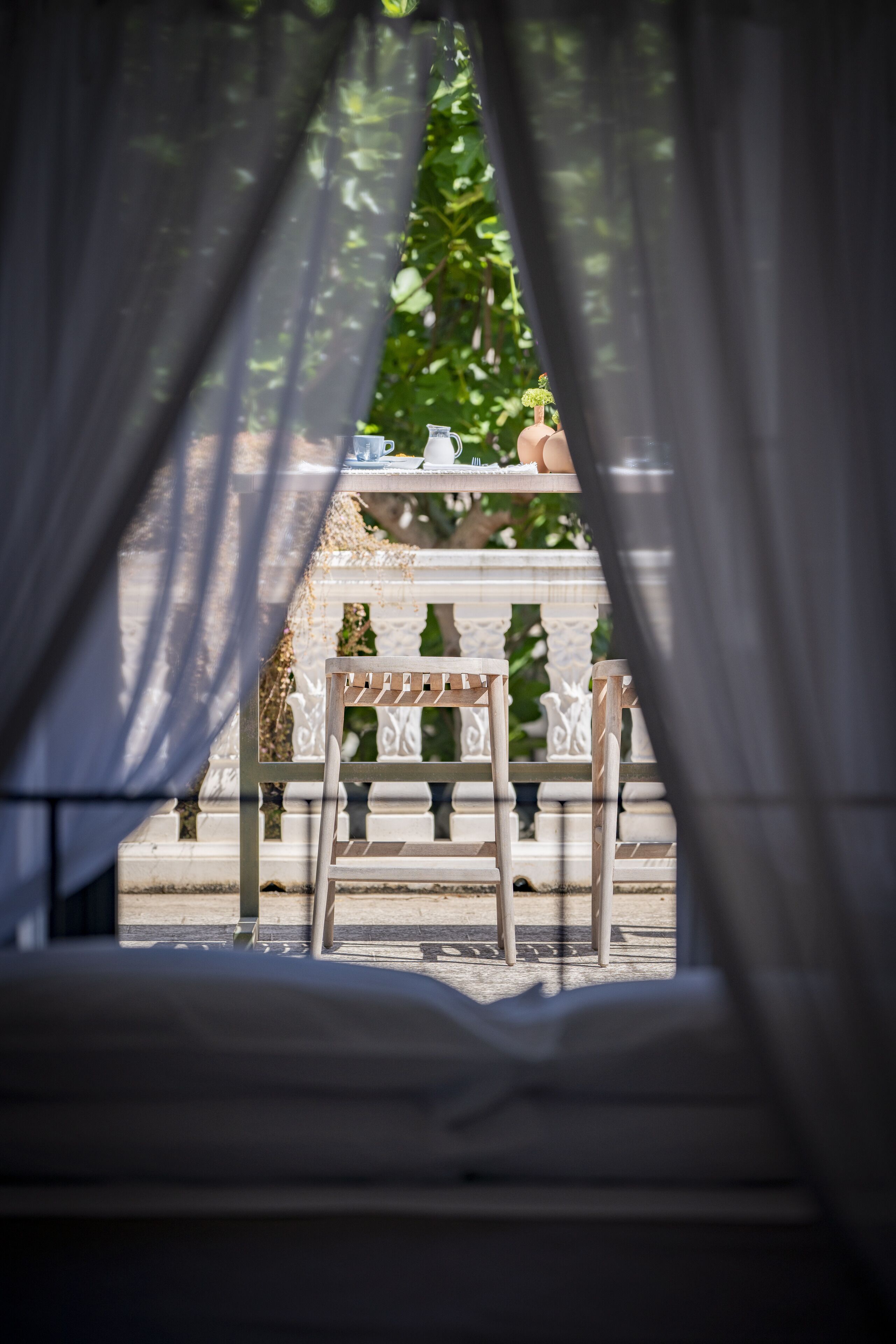 A view through sheer curtains of a stone balustrade and a small table with a chair, suggesting a balcony or terrace setting.