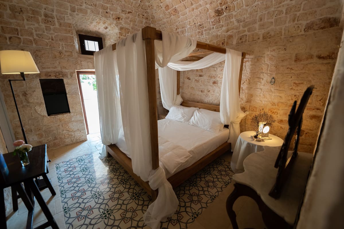 A four-poster bed with white linens in a room with exposed brick walls and patterned floor tiles.