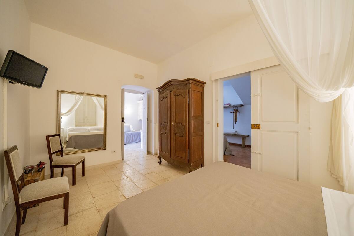 A bedroom with a bed, a wooden wardrobe, a chair, a television, and a mirror.