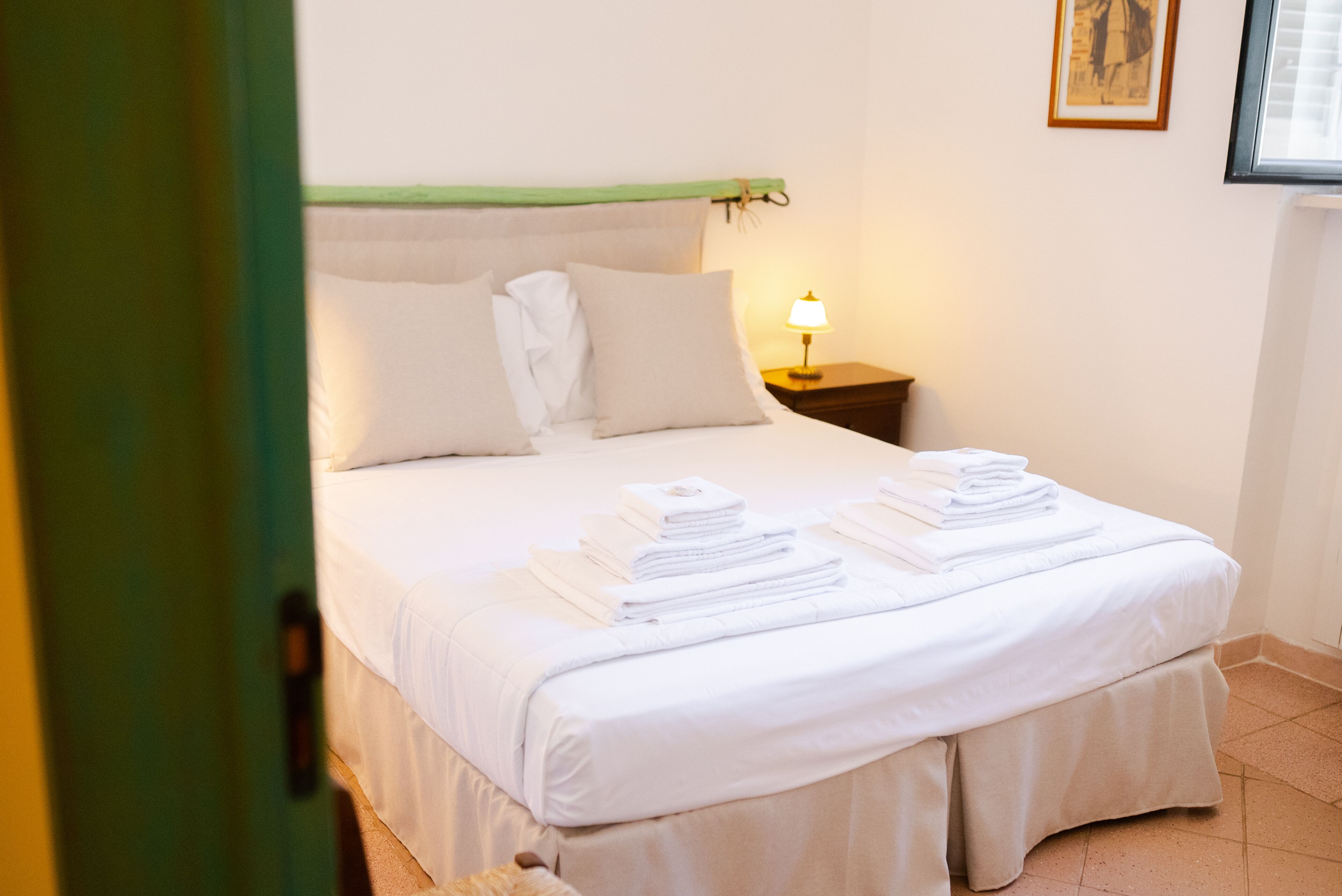 Una cama bien hecha con sábanas y toallas blancas, una lámpara de noche y un cuadro en la pared.