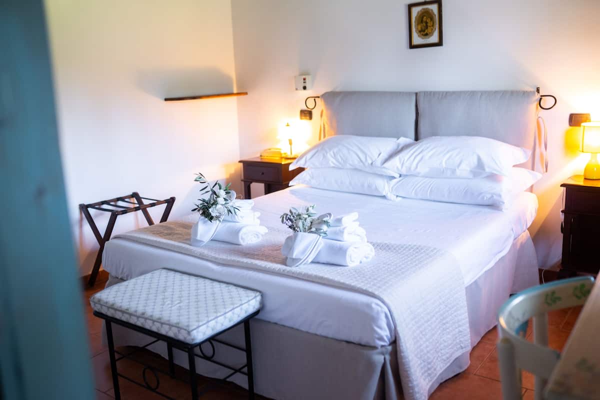 A neatly made bed with white linens, a bedside table with a lamp, and a chair with a cushion.