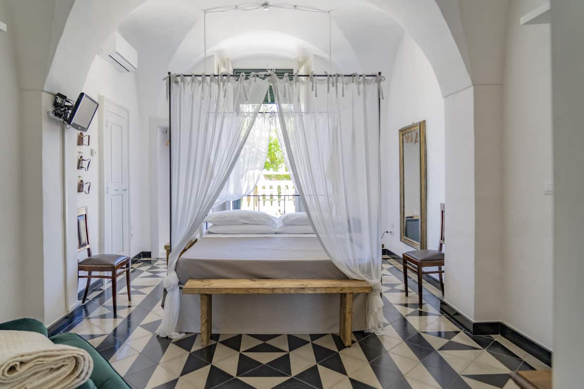 A bedroom with a bed, a bench, a chair, a mirror, and a view of the outside.