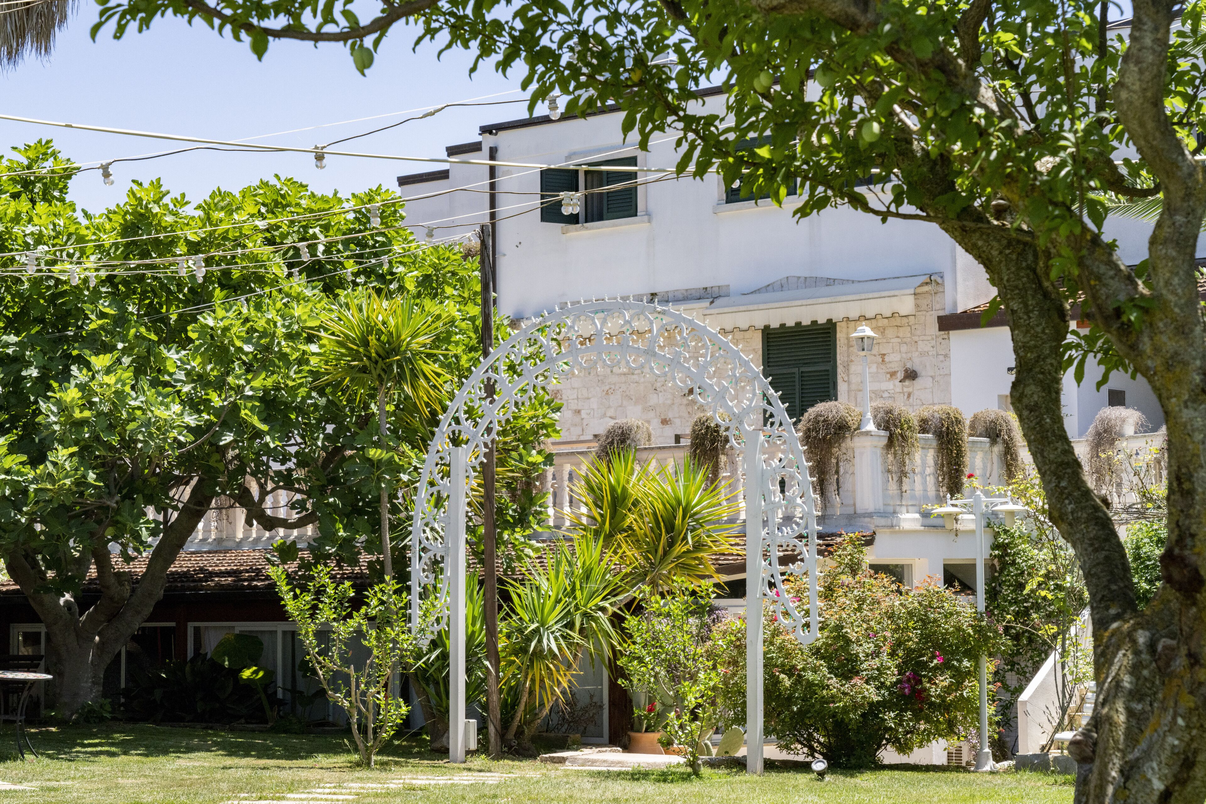 A garden with a white decorative archway, surrounded by lush greenery and a building with a tiled roof in the background.