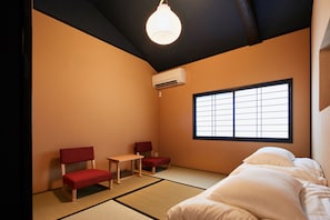 Una habitación con suelo de tatami, una cama con ropa de cama blanca, dos sillas rojas, una mesita y un ventanal con diseño de cuadrícula.