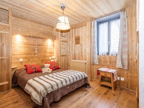 Ein Zimmer einer Holzhütte mit Bett, einem kleinen Tisch, einem Fenster mit Vorhängen und einer Heizung.
