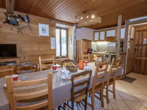 A wooden interior with a dining table set for four, surrounded by wooden chairs. A television is mounted on the wall, and a kitchen area is visible in the background.