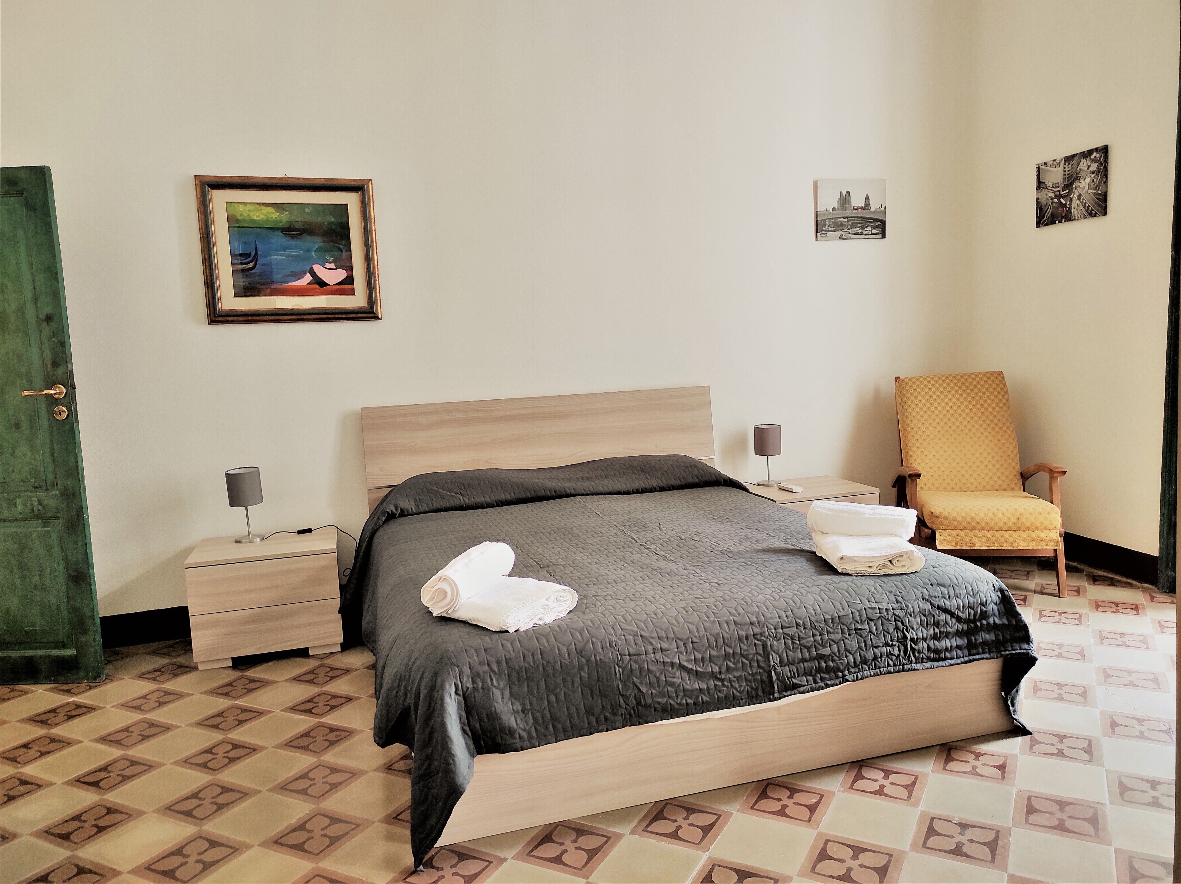 A neatly made bed with a dark quilt, two bedside tables with lamps, a chair, and artwork on the walls.