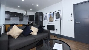A modern living room with a grey sofa, a glass coffee table, and a kitchen in the background. - New Oxford House Apartments (Liverpool)