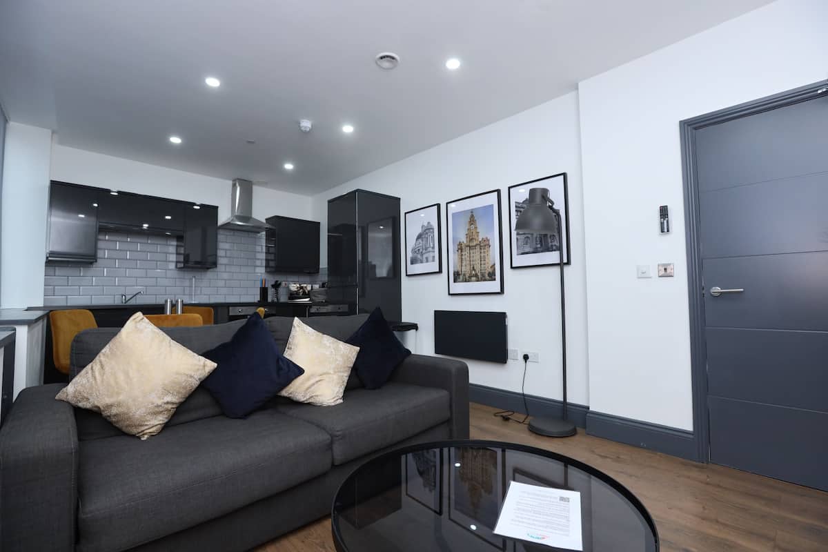 A modern living room with a grey sofa, a glass coffee table, and a kitchen in the background.