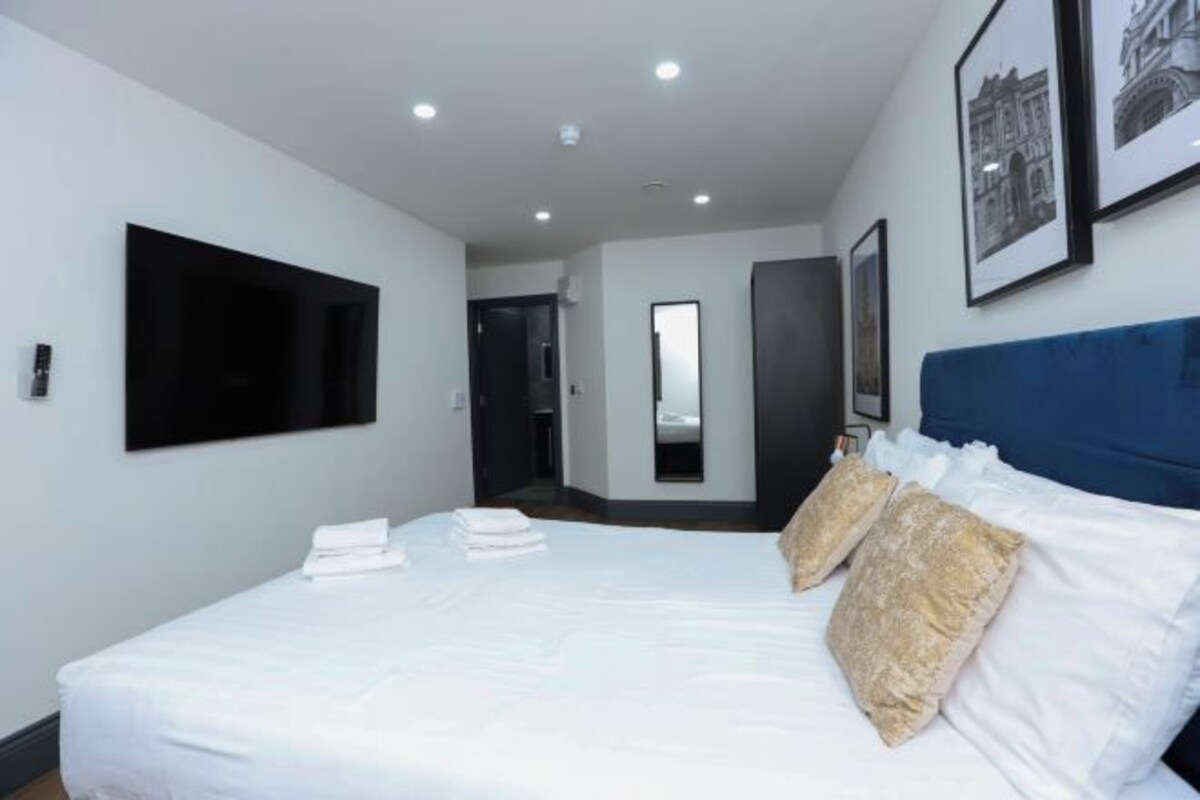 Una habitación de hotel moderna con una cama grande, un televisor de pantalla plana montado en la pared, un espejo y un cuadro enmarcado.