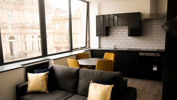 A modern apartment with a sofa, yellow chairs, a dining table, a kitchen with a black stove, and a view of a historic building. - New Oxford House Apartments (Liverpool)