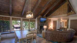 Ein geräumiges Wohnzimmer mit Kamin aus Stein, Holzbalkendecke, einem an der Wand befestigten Fernseher und einem Essbereich mit Tisch und Stühlen.