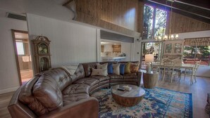 Ein geräumiges Wohnzimmer mit einer großen, ledernen L-Form-Couch, einem Couchtisch und einem gemusterten Teppich. Im Hintergrund sind eine Holz-Standuhr und ein Essbereich mit Tisch und Stühlen zu sehen.