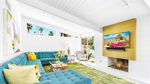 A modern living room with a blue sectional sofa, a flat-screen TV mounted on a gold wall, and a fireplace. - REGAL Palm Springs - Palm Springs Paradise: Your Private Poolside Retreat Awaits (Palm Springs)