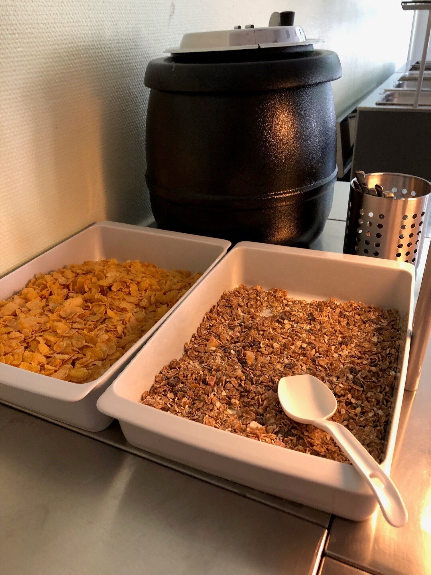 Zwei weiße Tabletts mit Frühstückscerealien, eines mit Cornflakes und das andere mit Granola, auf einem Tisch. Im Hintergrund steht ein schwarzer Topf.