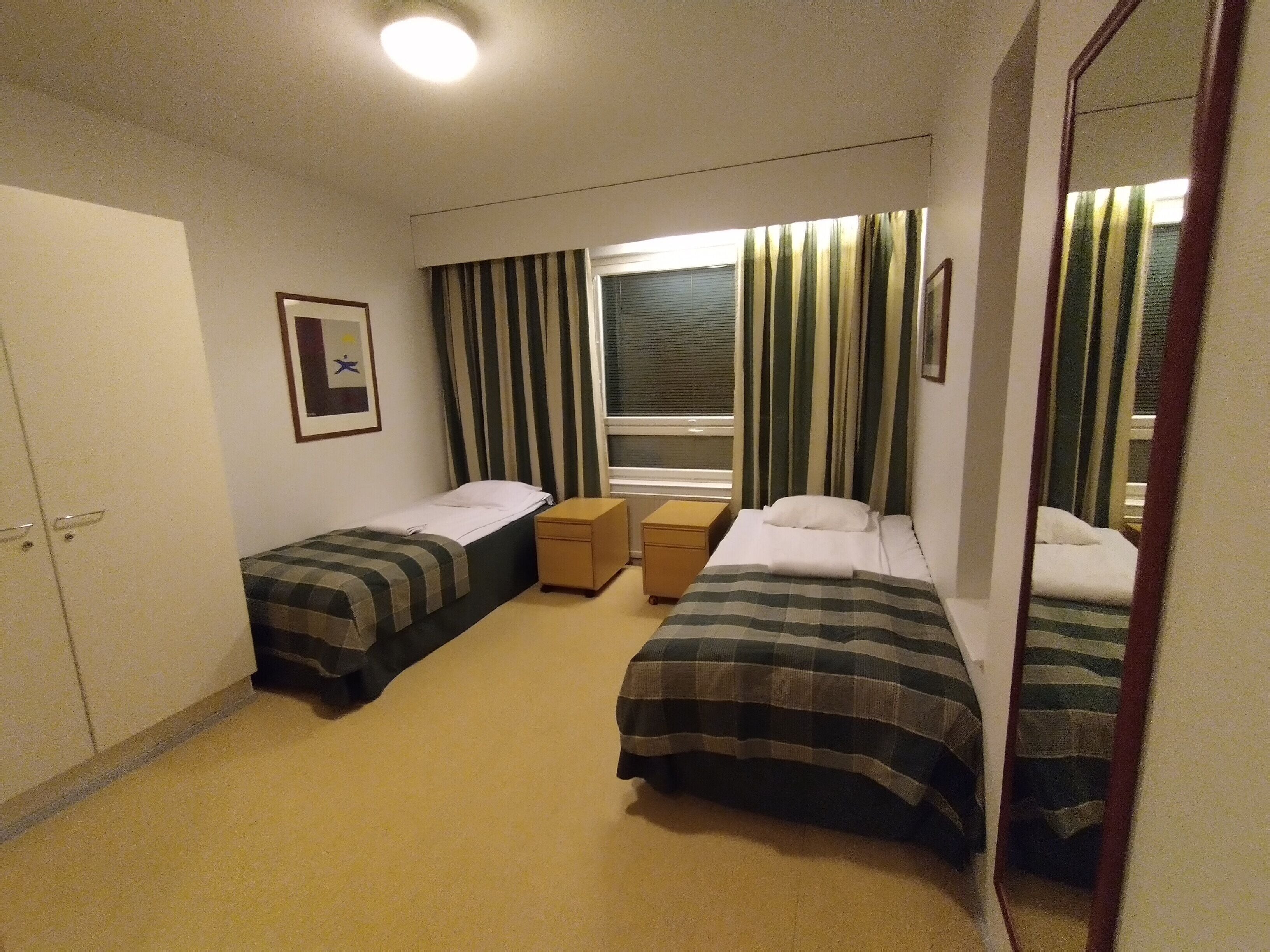 A hotel room with two single beds, a small wooden nightstand, a window with striped curtains, and a framed picture on the wall.