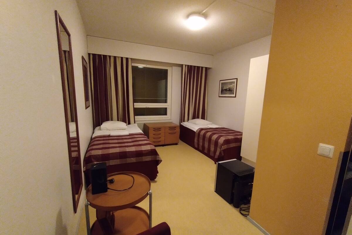 Ett kompakt hotellrum med två sängar, ett litet bord, en stol, ett skrivbord och ett fönster med gardiner.
