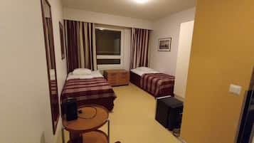 Habitación de hotel compacta con dos camas, una mesita, una silla, un escritorio y una ventana con cortinas.
