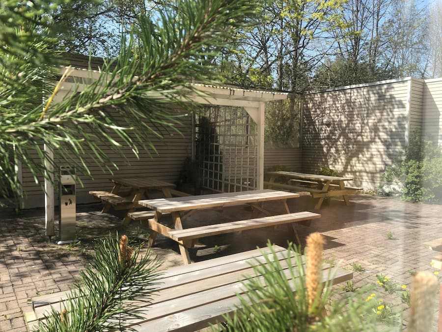 An outdoor patio area with wooden picnic tables and benches, surrounded by a brick floor and greenery.