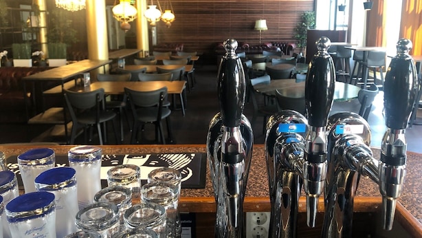 A restaurant interior with multiple tables and chairs, a bar area with taps, and a wooden wall in the background. - West Side Hotel (Turku)
